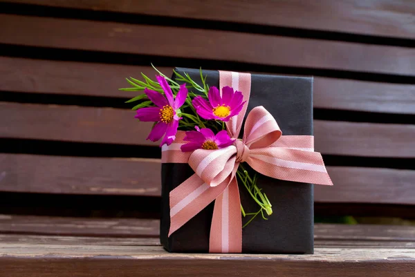 Black gift with pink ribbon and cosmea flowers on wooden background