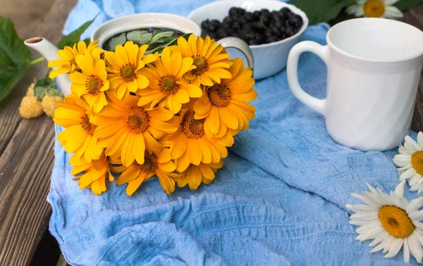 A small teapot in yellow daisies on a blue fabric along with black currants and yellow raspberries on a wooden background