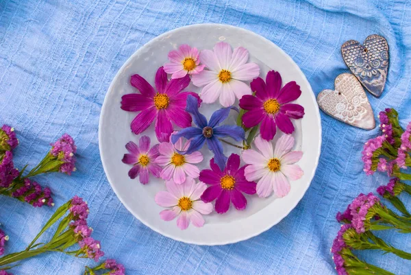 Cosmos and clitis flowers in a bowl of water, surrounded by flowers and ceramic hearts on a blue background