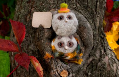 Two small owlets made of wool with beady eyes and a beak made of plastic in light colors with a hat and a scarf are sitting near a small hollow in a tree
