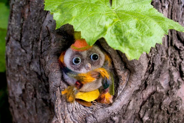 A toy small owl made of wool with beady eyes and a plastic beak in light colors sits near a small hollow in a scarf and hat under a grape leaf during the day