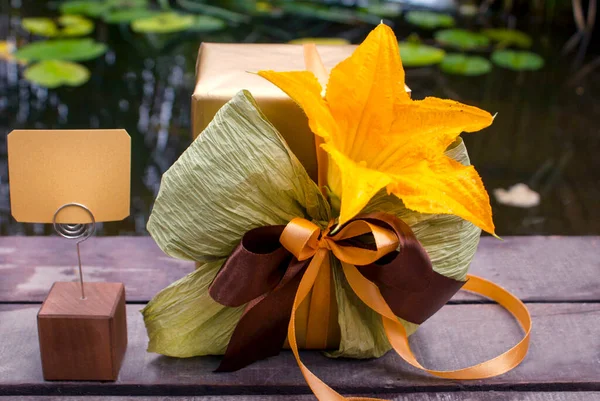 A golden gift with paper and textile ribbons, a zucchini flower and a wooden holder with a sign in the garden near the lake