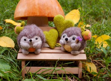 Date under a mushroom made of pumpkin and salt dough, on a plywood bench near two felted hedgehogs in the afternoon, surrounded by green grass, felted hearts and yellow leaves