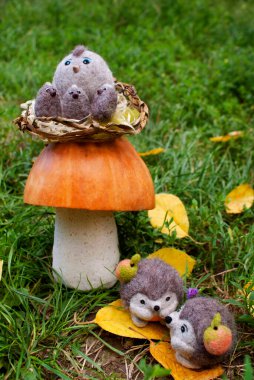 Mushroom made of pumpkin and salt dough, on which there is a nest with felted chicks and their mother, under it are two felted hedgehogs in the afternoon surrounded by green grass and yellow leaves