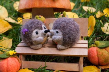 Date under a mushroom made of pumpkin and salt dough, on a plywood bench near two felted hedgehogs during the day, surrounded by green grass, felted pumpkins and yellow leaves