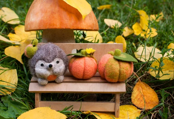 Felted hedgehog on a plywood bench in nature under a mushroom made of pumpkin and salt dough, outdoors during the day surrounded by felted pumpkins, green grass and yellow leaves