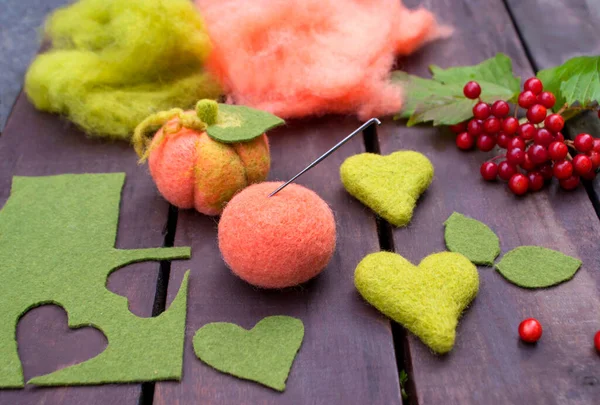 Pumpkin made using the dry felting technique, details and materials for making a felted pumpkin and two hearts made of wool on wooden boards surrounded by viburnum berries