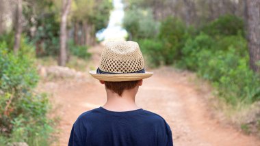 Boy with straw hat and dark blue t-shirt walking towards the beach between a forest of pine trees