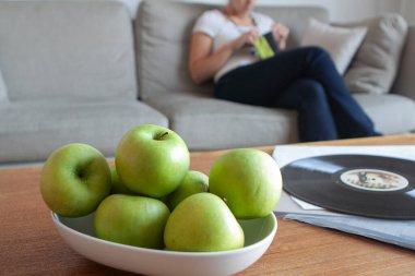 Fruit bowl of green apples on wooden table and unfocused woman sitting on the sofa in the background