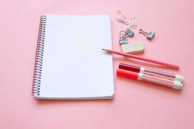 pink pencil, felt-tip pens and stationery items on notepad with blank page and on pink background