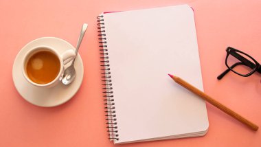 Blank page of a notebook next to a laptop and a white cup with coffee on a light pink background