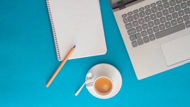 Blank page of a notebook next to a laptop and a white cup with coffee on a blue background