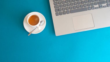 White cup with coffee and shiny metal spoon next to laptop on blue background