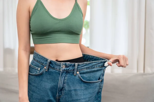 Sporty woman is smiling while wearing in loose big jean to showing successfully of weight loss after healthy diet and hard exercise in living room.