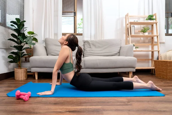 Young woman is practicing yoga with cobra pose during exercise on mat in her living room at home.