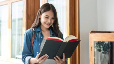 Young woman student is standing near the window and reading literature to relaxation while taking a break between lesson classes in college.