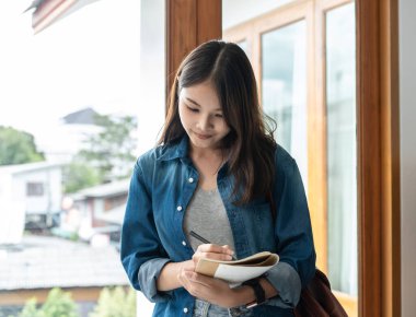 Young woman student is standing near the window to writing schedule and taking notes on notebook while taking a break between lesson classes in college.