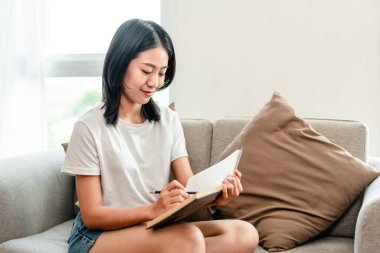 Half-Japanese woman is writing a diary on the sofa in her living room, Daily routine record, Relaxing time on vacation, Happy time, Living at home, Comfortable corner.