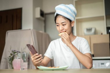 Woman is having breakfast with crackers mixed with various grains and using mobile phone to update her morning news, Small room in condominium background, Wake-up activities.