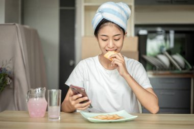 Woman is having breakfast with crackers mixed with various grains and using mobile phone to update her morning news, Small room in condominium background, Wake-up activities.