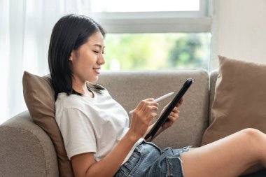 Half-Japanese woman sitting on the sofa using tablet while on vacation, Rest at home, Living room, Relax time, Touch screen tablet, Go on internet, Favorite corner, Personal space.