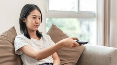 Half-Japanese woman turns on the TV to watch a movie or drama in the living room, Hold the remote to turn on-off the TV, Relaxing time on vacation, happy corner, Remote control.