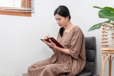 Beautiful woman is writing notes and work schedule in notebook while sitting to relaxation on carpet in the living room.