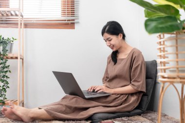 Beautiful woman is using laptop to chatting and talking with friends on video call while sitting to relaxation on carpet in the living room.
