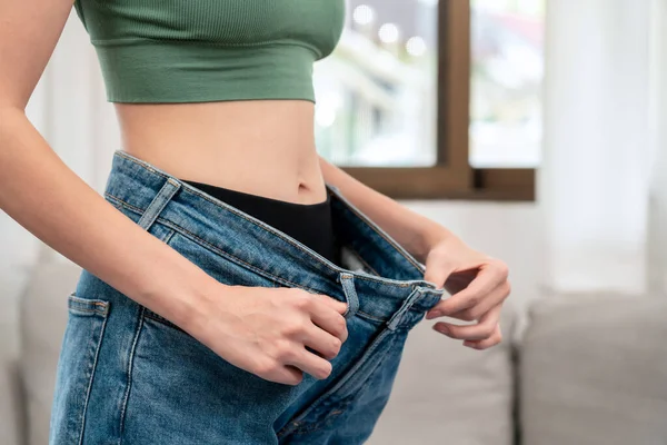 Sporty woman is smiling while wearing in loose big jean to showing successfully of weight loss after healthy diet and hard exercise in living room.
