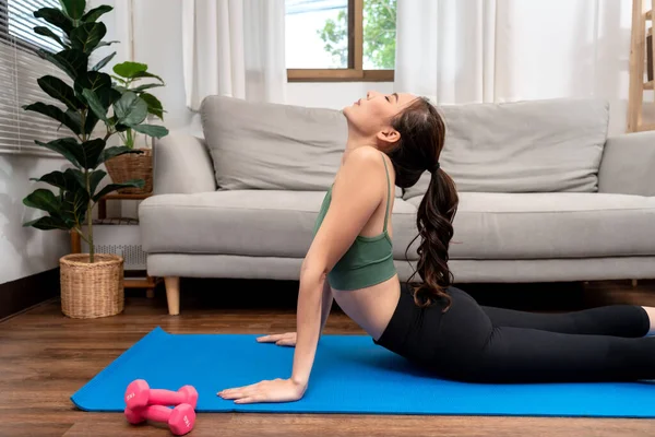 Young woman is practicing yoga with cobra pose during exercise on mat in her living room at home.