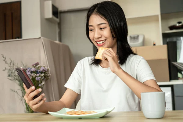 Ασιάτισσα Τρώει Κράκερ Για Πρωινό Και Χρησιμοποιεί Κινητό Της Τηλέφωνο — Φωτογραφία Αρχείου