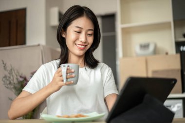 Asian woman drinking hot cocoa with crackers mixed with various grains and using tablets to update her morning news, Small room in condominium background, Wake-up activities.