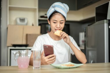 Woman is having breakfast with crackers mixed with various grains and using mobile phone to update her morning news, Small room in condominium background, Wake-up activities.