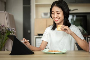 Woman is having breakfast with crackers mixed with various grains and using tablets to update her morning news, Small room in condominium background, Wake-up activities.
