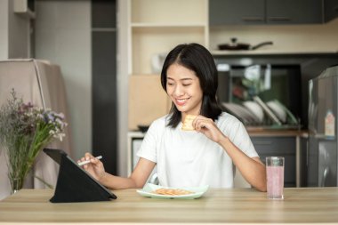 Woman is having breakfast with crackers mixed with various grains and using tablets to update her morning news, Small room in condominium background, Wake-up activities.