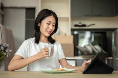 Asian woman drinking hot cocoa with crackers mixed with various grains and using tablets to update her morning news, Small room in condominium background, Wake-up activities.