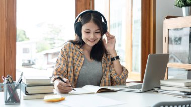 Young woman student is taking notes in a notebook while learning lessons online with teacher and doing homework assignment after finished class in college.