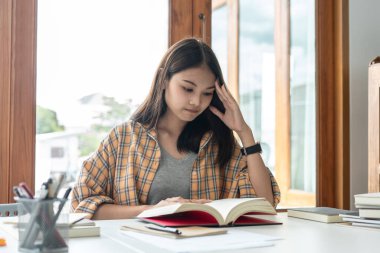 Young woman student is suffering serious tiredness and headaches while reading hard textbook to preparing for examination in college.