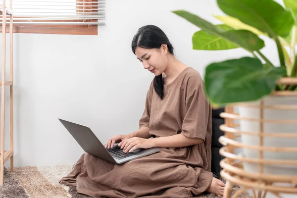 Beautiful woman is using laptop to chatting and talking with friends on video call while sitting to relaxation on carpet in the living room.