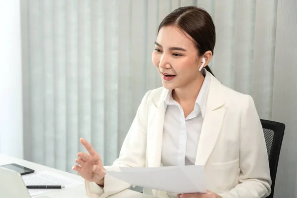 Asian businesswoman talking on the wireless headphones with a colleague in a private office, Telephone communication, Emote conversation, Modern office with laptop and documents on the desk.