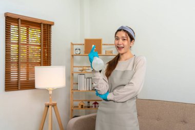 Housekeeper is wearing protective gloves and holding cleanser bottle to prepare wiping dirt in living room while working to perform routine house cleaning.