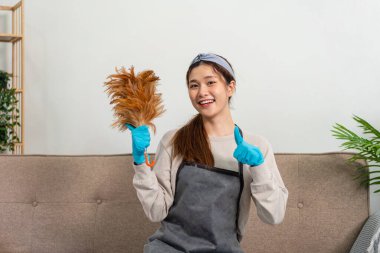Housekeeper is wearing protective gloves and holding feather duster to showing happy thumbs up gesture after dusting dirt and working to perform routine house cleaning.