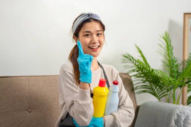 Housekeeper is wearing protective gloves to holding cleanser bottle and smiling to showing happy thumbs up gesture after successfully working to perform routine house cleaning.