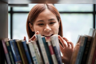 Female student is standing in front of the bookshelf to choosing textbook or literature in library between take a break from studying lessons class at university.