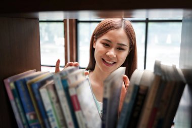 Female student is standing in front of the bookshelf to choosing textbook or literature in library between take a break from studying lessons class at university.