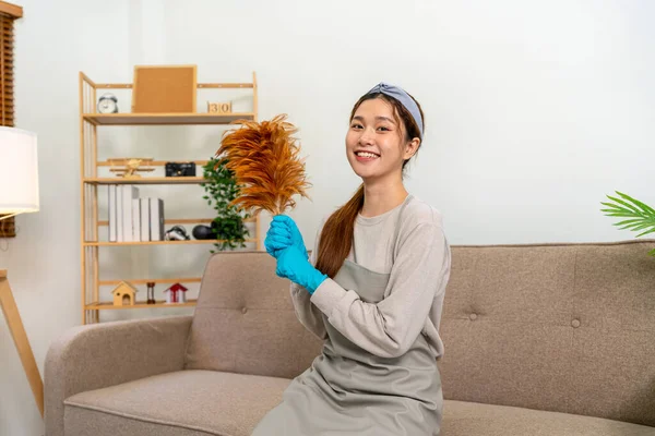 Housekeeper is wearing protective gloves and holding feather duster to cleanups and dusting in living room while working to perform routine house cleaning.
