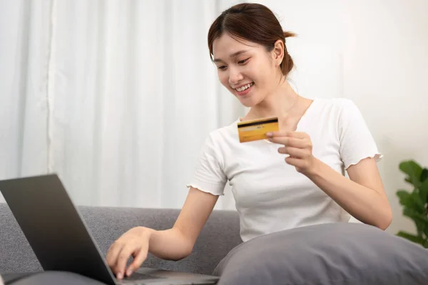 Teenage girl is holding credit card and typing credit card number for payment after shopping online on laptop while sitting to relax comfortable on the couch in living room.