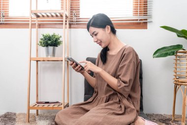 Beautiful woman is using smartphone to chatting with friends and playing games while sitting to relaxation on carpet in the living room.