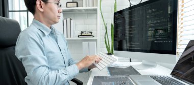 Male programmer is programming to develop program app and website of corporate while working to checking program code and debugging on multiple screen at modern software office.