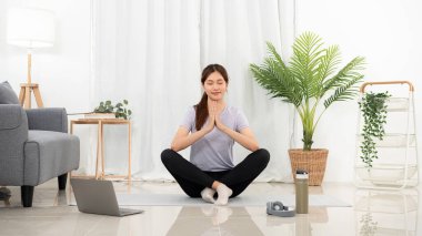 Young woman is relaxing into the lotus position for meditation on mat while taking yoga online class and practicing yoga in her living room at home.